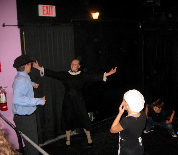 A smiling actor with arms spread out wide, while other actors watch in shock